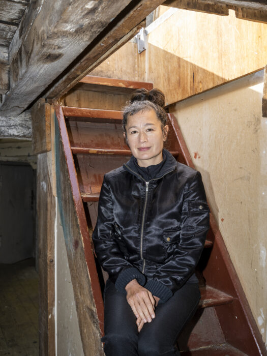 A woman with brown hair pulled into a bun wearing a black zip up jacket, sitting on a staircase