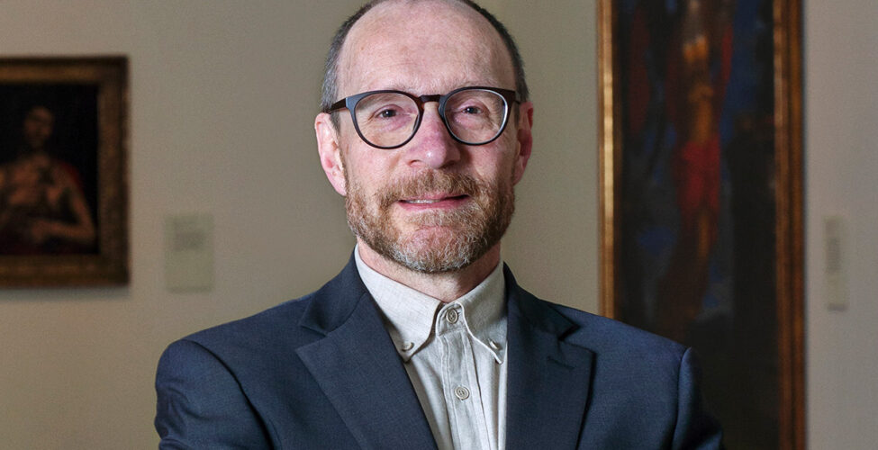 Photo of a man wearing glasses, a collared shirt and jacket, with his hands crossed in front of him.
