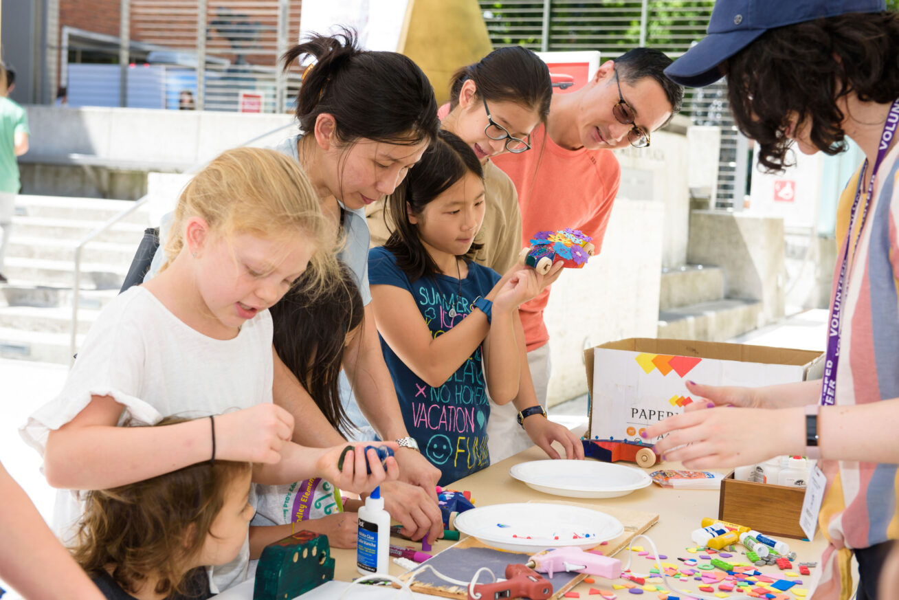 Photo of kids and parents at an artmaking table, covered in glue guns, bottles of glue, and other art supplies